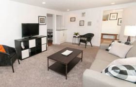 Spacious living room with modern furniture, a flat-screen TV, and a cozy corner with a wicker chair and small table.
