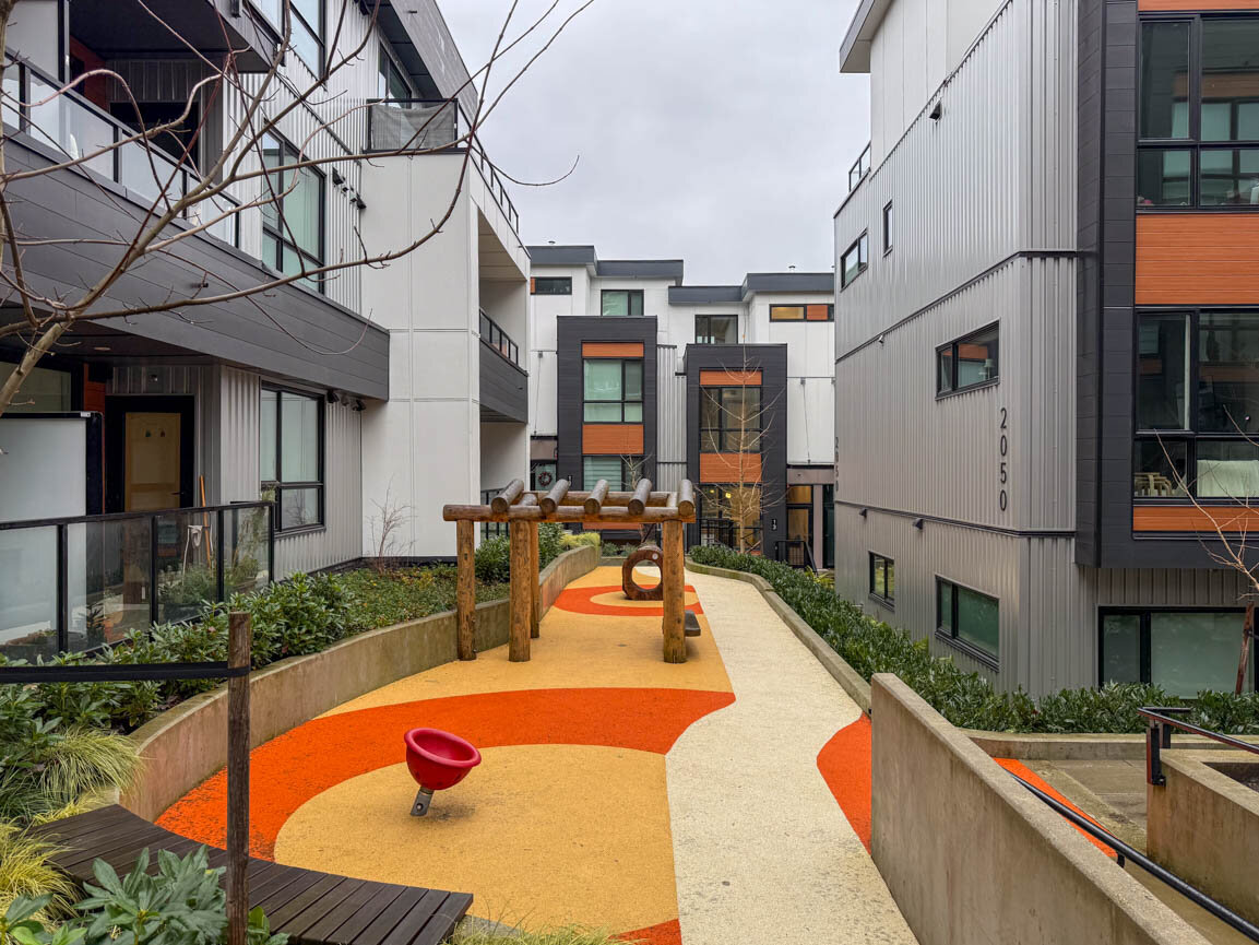 Outdoor playground courtyard with colorful rubber surfacing, a wooden log climber, and a red bowl swing. - Hendry Place