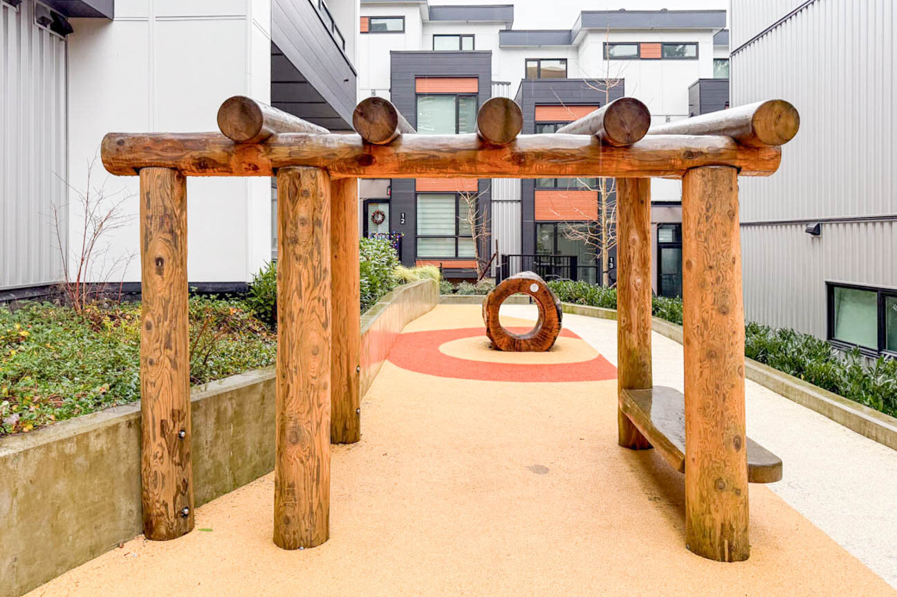 Outdoor play area with a rustic log climbing frame and circular wooden ring on a rubberized surface. - Hendry Place