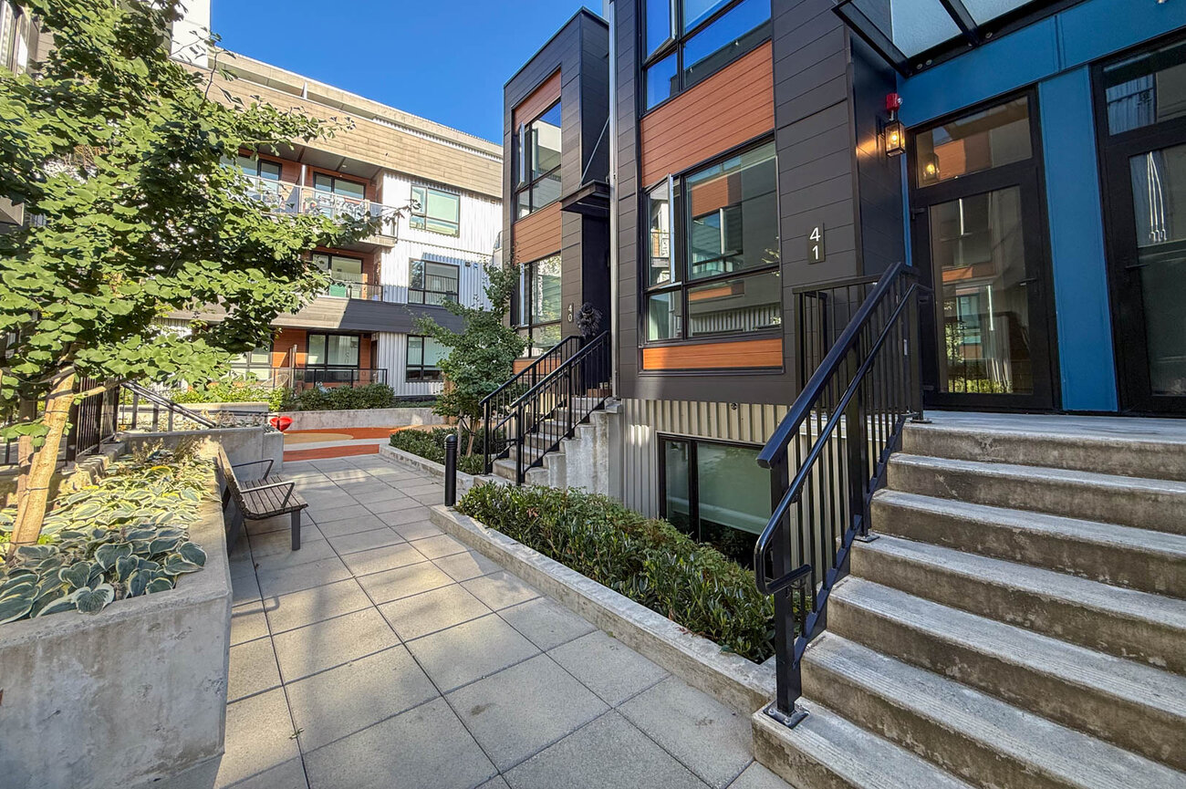 Courtyard entry featuring concrete pavers, landscaped planters, and a modern multi-color facade with black stair railings. - Hendry Place