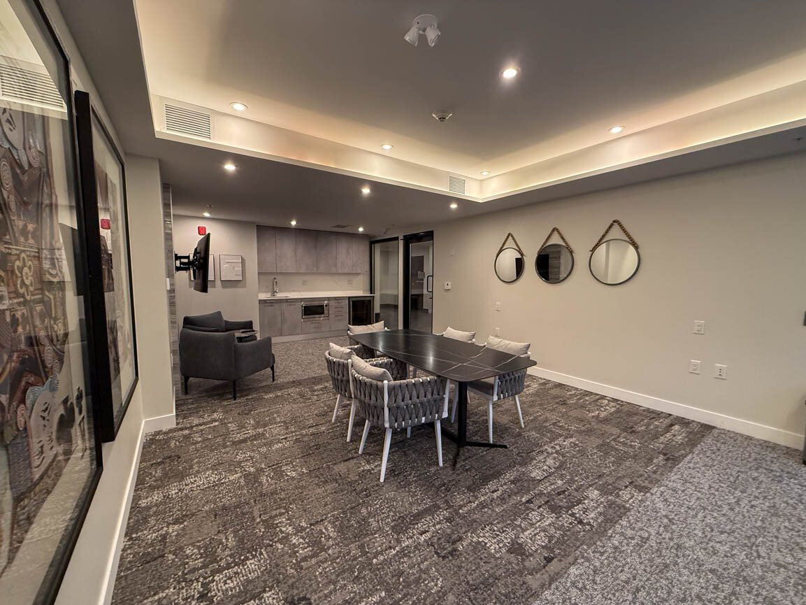 Shared lounge with dark dining table, woven chairs, gray carpet, and a kitchenette with built-in oven. - Hendry Place