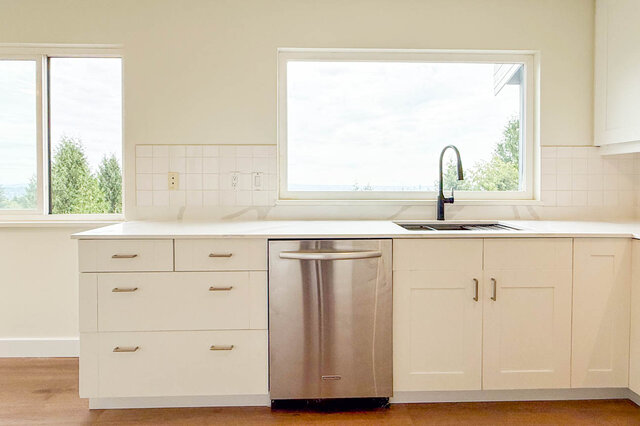 Bright kitchen with white cabinetry, white countertops, stainless dishwasher, and hardwood floors beneath two windows.