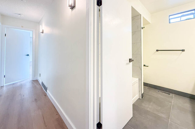 Open hallway with light hardwood floors and wall sconces leading to a tiled bathroom with a white vanity. - 1350 Crestwell Road, West Vancouver