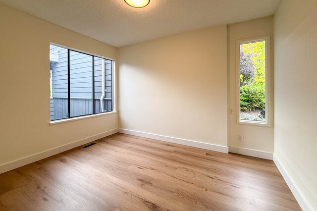 Empty bedroom with light hardwood floors and two windows framing a view of greenery and a neighboring home. - 1350 Crestwell Road, West Vancouver