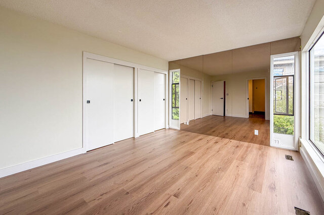 Bright bedroom with light hardwood floors, white sliding closet doors, and a mirrored wall reflecting greenery outside.
