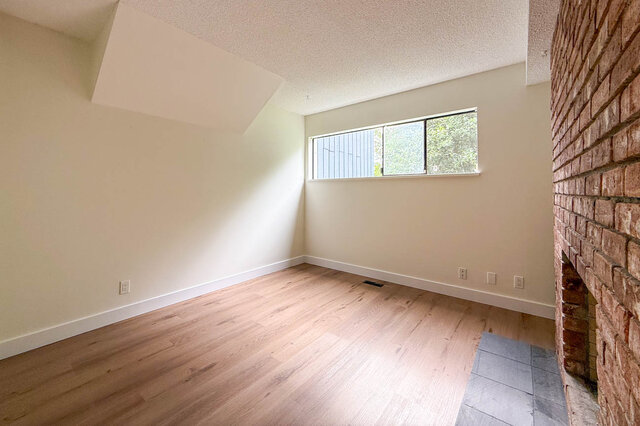 Empty living room with light hardwood floors and a red brick fireplace, plus a horizontal window framing greenery.