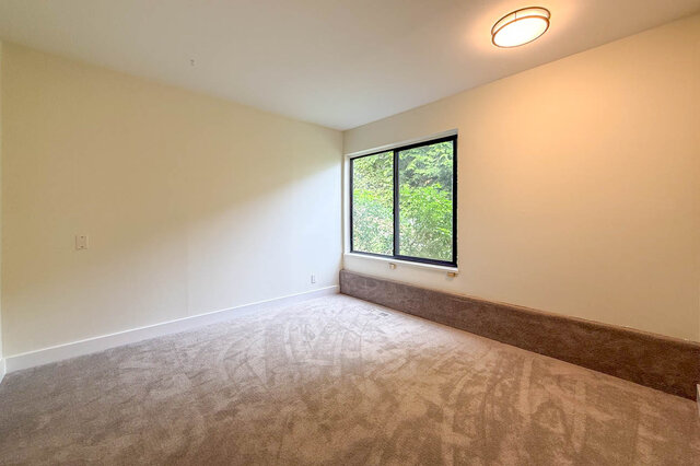 Empty carpeted room with a window overlooking lush green trees and a warm ceiling light. - 1350 Crestwell Road, West Vancouver