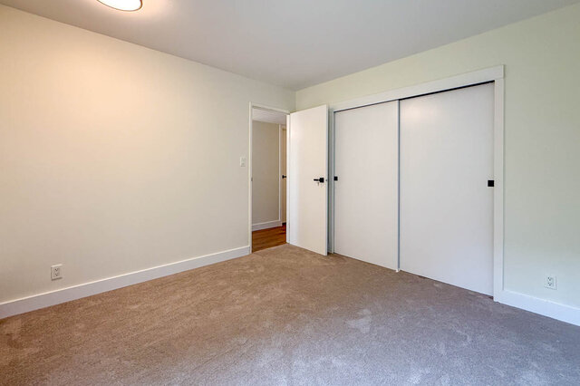 Empty bedroom with beige carpet, white sliding closet doors, and an open doorway revealing hardwood floors beyond.