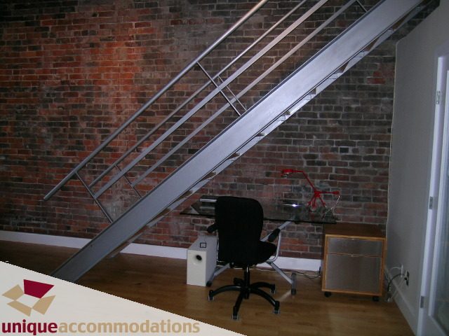 Bright living room with exposed brick wall, modern staircase, and hardwood floors. - PH3  603 - 1155 Mainland, Vancouver Downtown