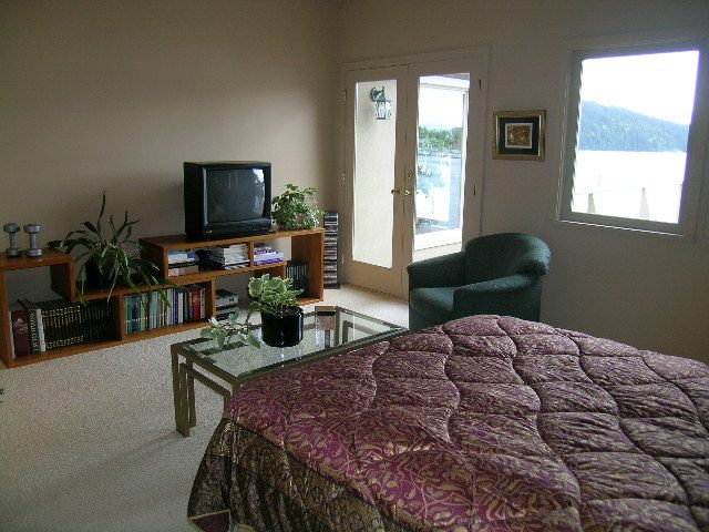 Bright bedroom with purple quilt, wooden bookshelf, and view of water outside. - 4527 Stonehaven Avenue, North Vancouver
