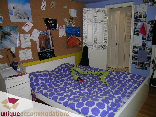 Furnished bedroom with blue and white patterned bedding, corkboard wall, and open door leading to another room. - 3664 Creery Ave., West Vancouver