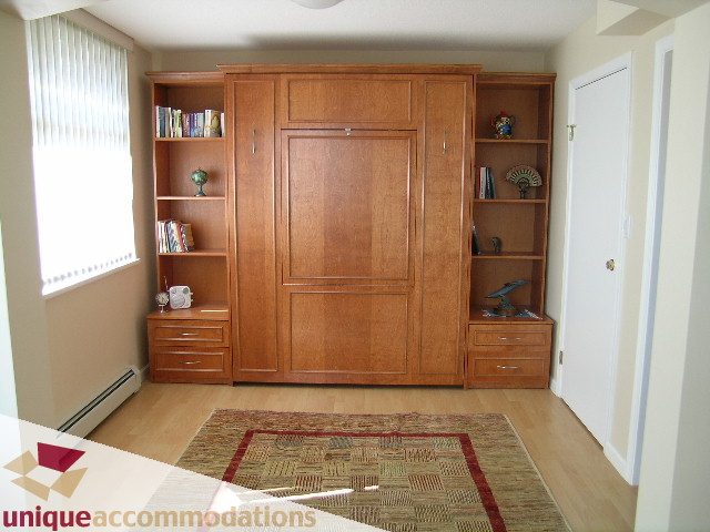 Bright living room with wooden built-in shelving and desk, hardwood floors, and a patterned rug. - 322 - 1228 Marinaside Crescent, Vancouver Downtown