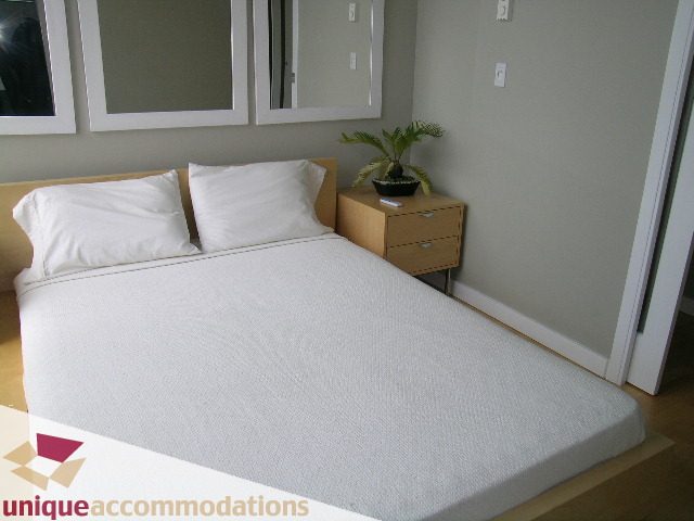 Furnished bedroom with white bedding, wooden headboard, and small nightstand with plant. - 2502 - 1077 Marinaside Crescent, Vancouver Downtown