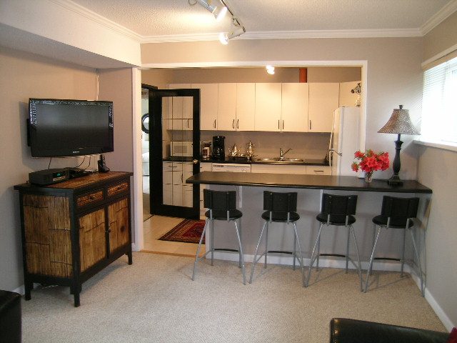 Bright kitchen with black countertops and bar stools, open to living area with TV and lamp. - 470 Ellis Street, North Vancouver