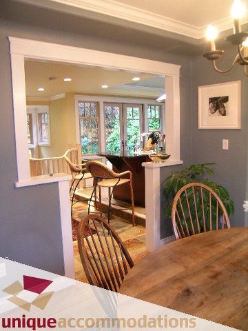 Dining area with wooden table and chairs, leading to a kitchen with large windows and greenery. - 4497 Marine Dr., West Vancouver