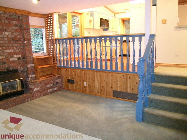 Bright living room with fireplace, wooden staircase, and kitchen in the background. - 4655 Clovelly Walk, West Vancouver