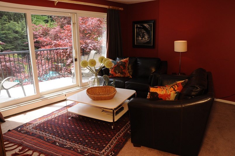 Spacious living room with red walls, black leather couch, and large windows showing vibrant foliage. - Guest House - 440 Stevens Drive, West Vancouver