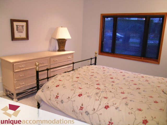Furnished bedroom with floral bedding and a dresser, large window with blinds. - 3967 West Broadway, Vancouver West Side