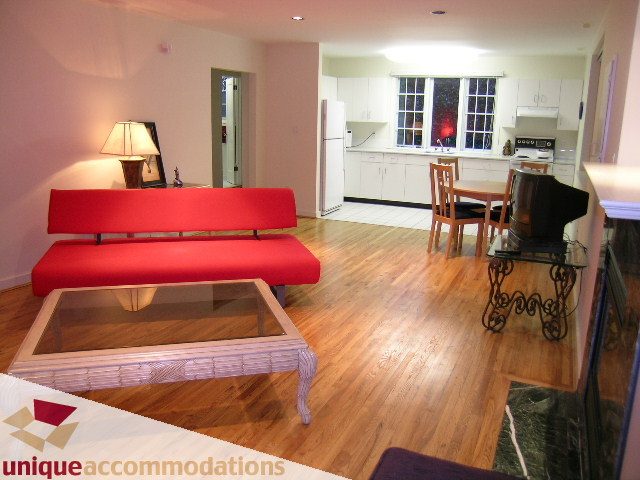Bright living room with red couch, hardwood floors, and a kitchen in the background. - 3967 West Broadway, Vancouver West Side