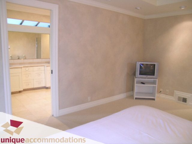 Furnished bedroom with a flat-screen TV and a view into a bathroom with a sink and mirror. - 6189 Nelson Avenue, West Vancouver