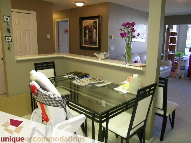 Furnished dining area with a glass table and white chairs, leading to a kitchen with a bar counter and vase of flowers.