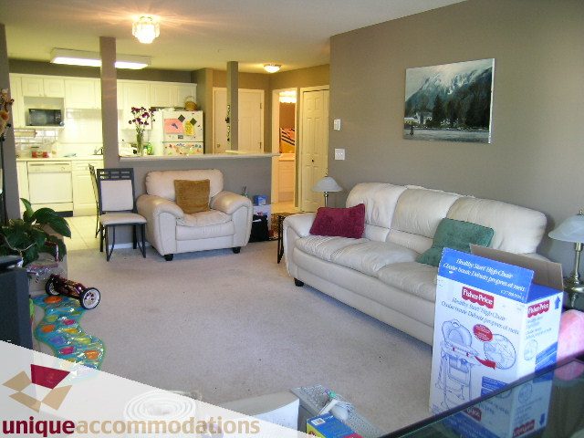 Furnished living room with white furniture, kitchen in background, and artwork on wall. - 314 - 855 W. 16th Street, North Vancouver