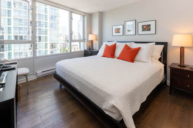 Bedroom with city view and hardwood floors, featuring a neatly made bed with orange accent pillows.