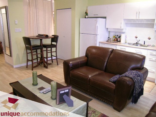Bright kitchen with white cabinets and a white refrigerator, open to a living area with brown leather furniture and...
