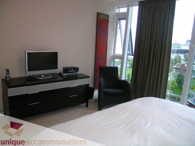 Spacious bedroom with a large window offering a city view, modern furniture, and a flat-screen TV on a sleek black console.