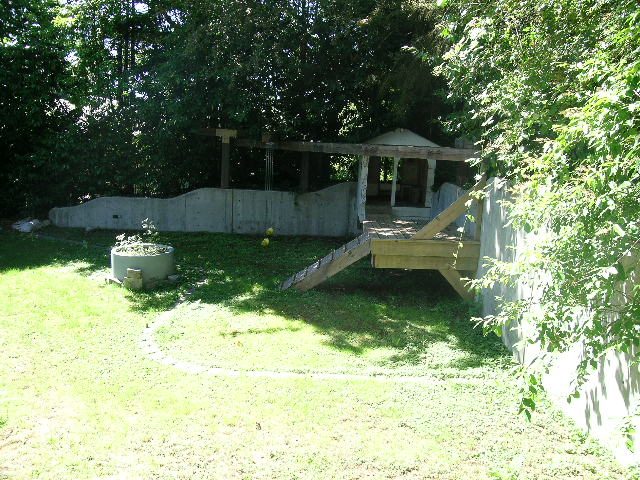 Furnished backyard with wooden deck and concrete walls, surrounded by lush greenery. - 947 Huntingdon Crescent, North Vancouver