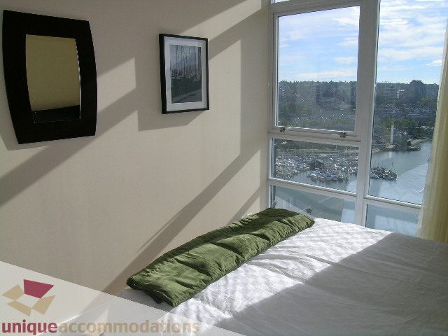 Spacious bedroom with a large window offering a stunning city view, featuring a neatly made bed with green accents and a...