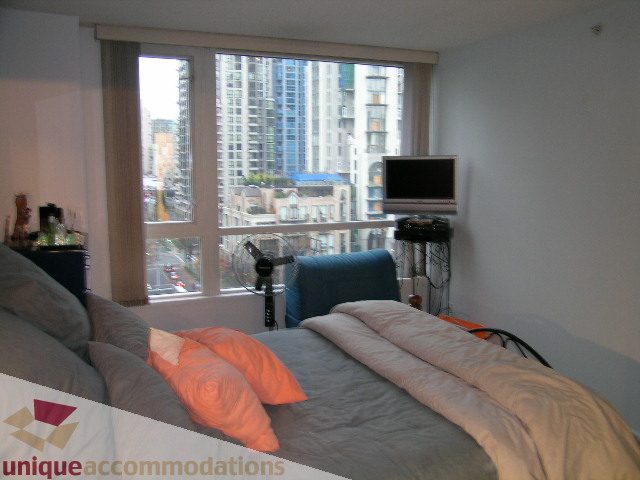 Spacious bedroom with a cozy bed, orange pillows, and a cityscape view from the large window. - 1306-388 Drake St, Vancouver Downtown