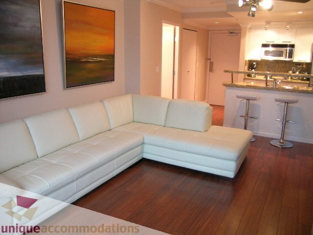 Furnished living room with white leather sectional, hardwood floors, and kitchen in the background.