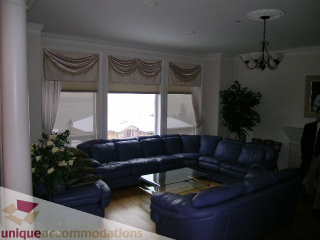 Furnished living room with blue leather couches, hardwood floors, and large windows with draped curtains. - 1659 Langton Place, West Vancouver