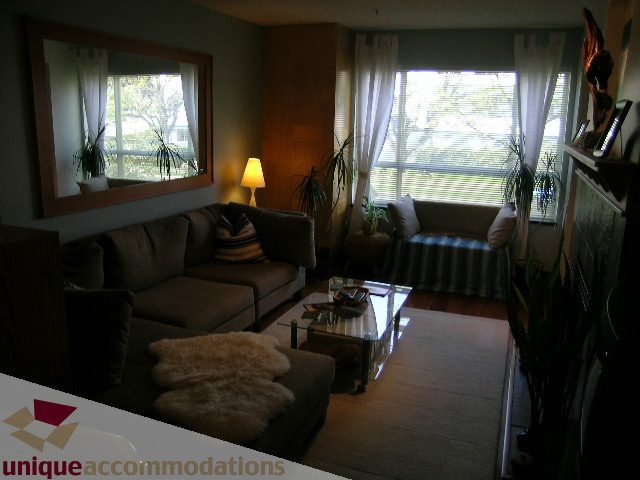 Furnished living room with large windows, cozy seating, and a wooden accent wall. - 201-365 E 1st Ave, North Vancouver