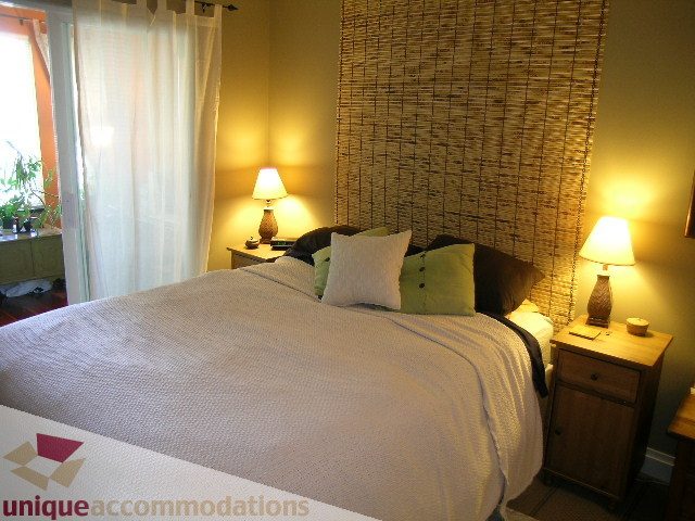 Furnished bedroom with cozy bedding, bamboo headboard, and bedside lamps. - 201-365 E 1st Ave, North Vancouver