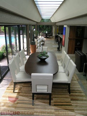 Furnished dining area with modern white chairs and a large table, leading to a glass-enclosed patio with a pool view. - 455 Keith Road, West Vancouver