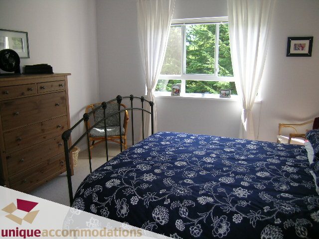 Bright bedroom with large window, blue floral bedding, and wooden furniture. - 304 - 1281 Parkgate Avenue, North Vancouver