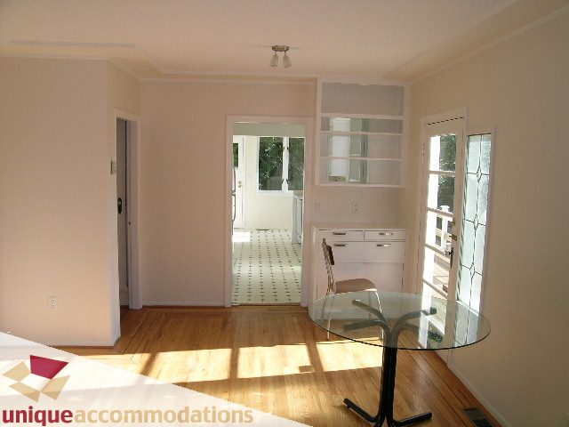 Bright living room with hardwood floors and a glass table, leading to a kitchen with white cabinets and a city view.