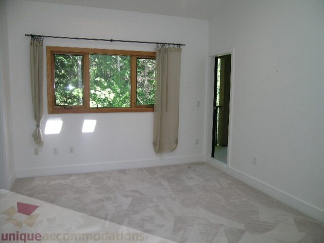 Bright bedroom with large windows and light-colored walls, featuring a partial view of a door leading to another room.