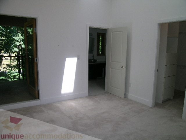 Bright living room with white walls, open door leading to bathroom, and large windows with a view of greenery. - 4215 Evergreen Ave, West Vancouver
