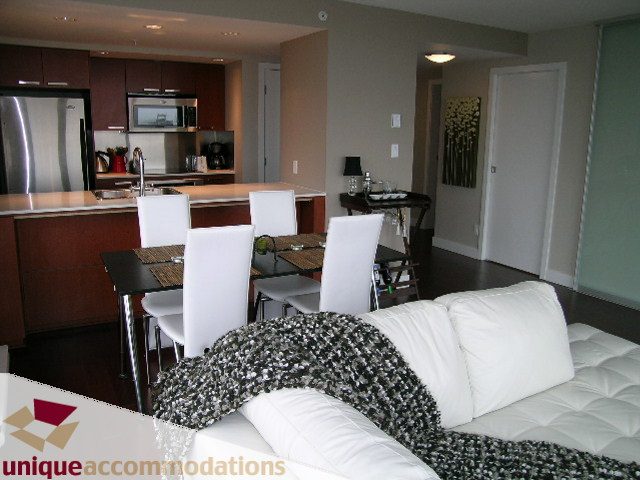 Modern kitchen with stainless steel appliances and wooden cabinets, adjacent to a cozy living area with white furniture...