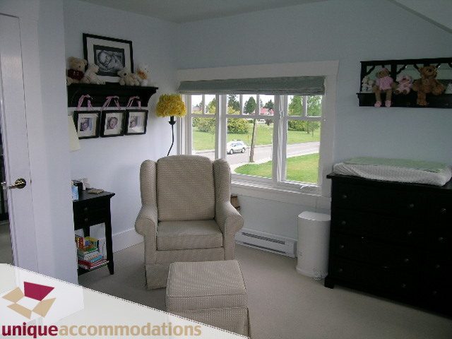 Bright living room with a view of the street and greenery, featuring a cozy armchair and a dresser. - 1505 Grand Blvd., North Vancouver