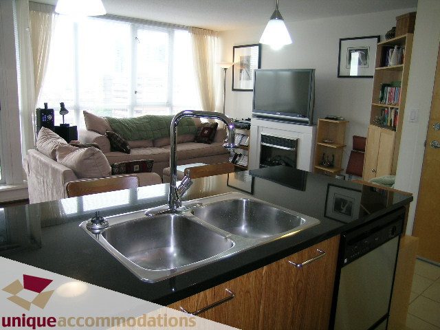 Bright kitchen with stainless steel sink and black countertops, adjacent to a cozy living area with a sofa and large windows.