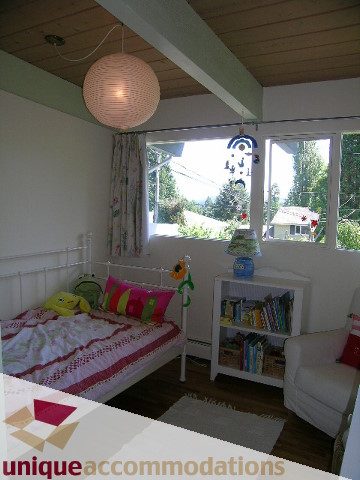Bright bedroom with hardwood floors, colorful bedding, and a large window offering a view of the outdoors. - 2556 Belloc Street, North Vancouver