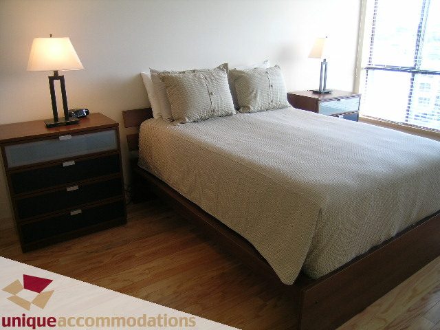 Furnished 2-bedroom condo with hardwood floors, modern nightstands, and a large window with city view.
