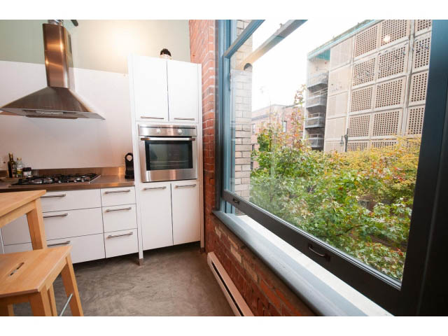 Bright kitchen with white cabinets, stainless steel appliances, and a city view through large windows. - 210 - 55 E Cordova St, Vancouver Downtown