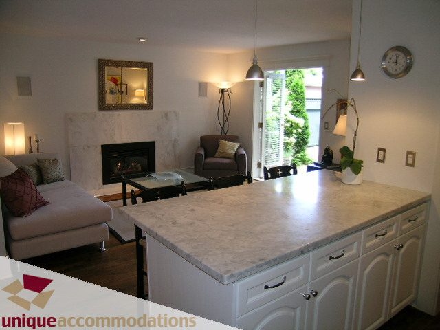 Bright kitchen with white cabinets and marble countertops, adjacent to a cozy living room with a fireplace and modern decor.