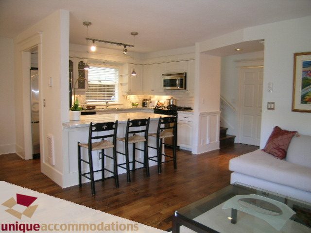 Bright kitchen with white cabinetry and hardwood floors, leading to a cozy living area with a white couch and glass...