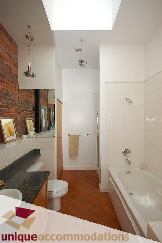 Bright bathroom with white fixtures, wooden floors, and a brick accent wall. - PH1-1178 Hamilton Street, Vancouver Downtown